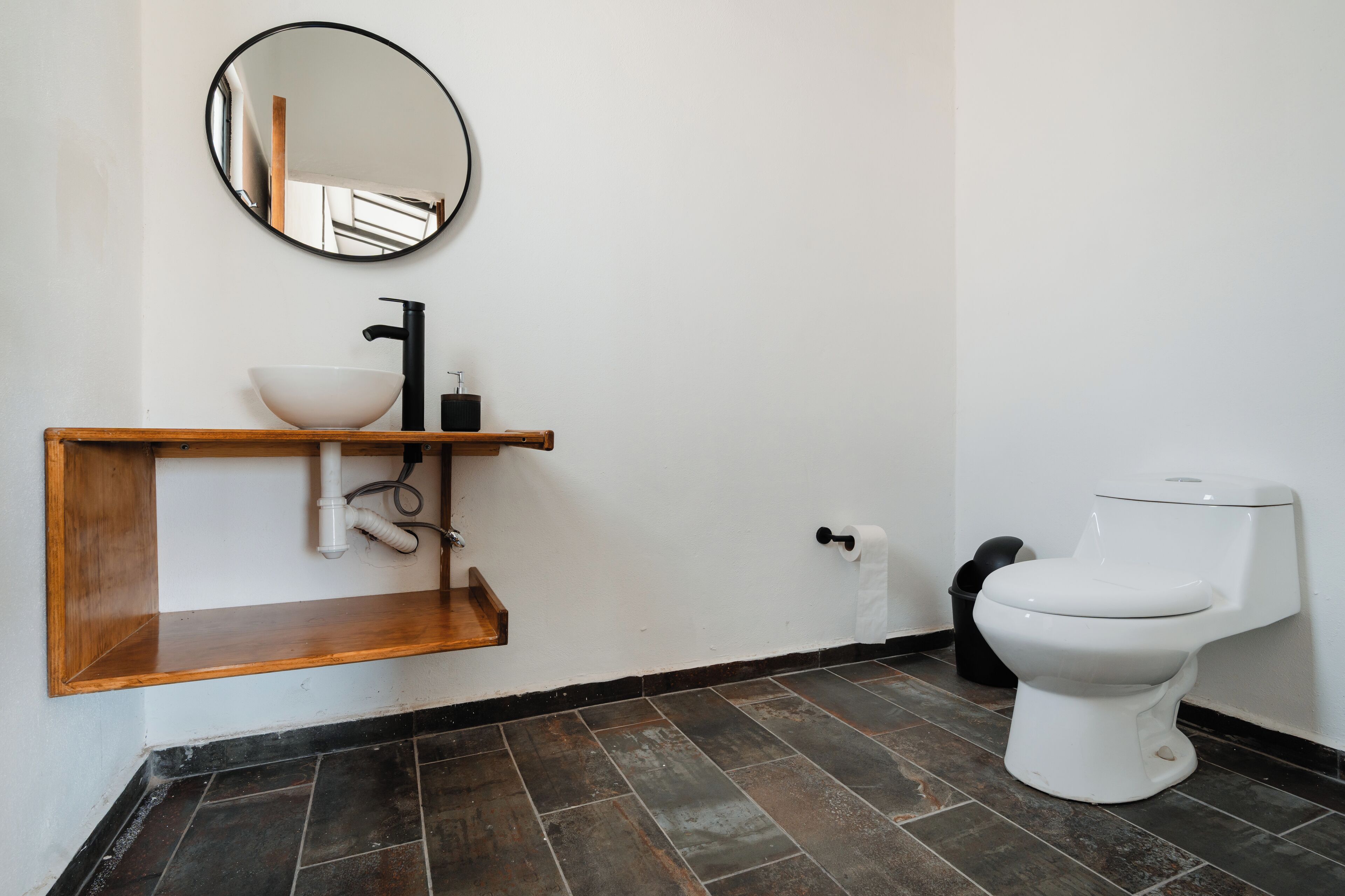 Ein Badezimmer mit einem Holzkosmetikschrank, einem runden Spiegel, einem weißen Waschbecken, einem schwarzen Wasserhahn, einer weißen Toilette und dunklen Fliesenböden.