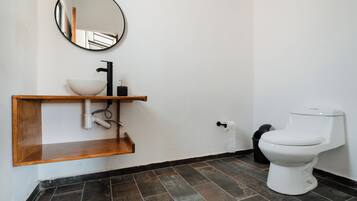 Ein Badezimmer mit einem Holzkosmetikschrank, einem runden Spiegel, einem weißen Waschbecken, einem schwarzen Wasserhahn, einer weißen Toilette und dunklen Fliesenböden.