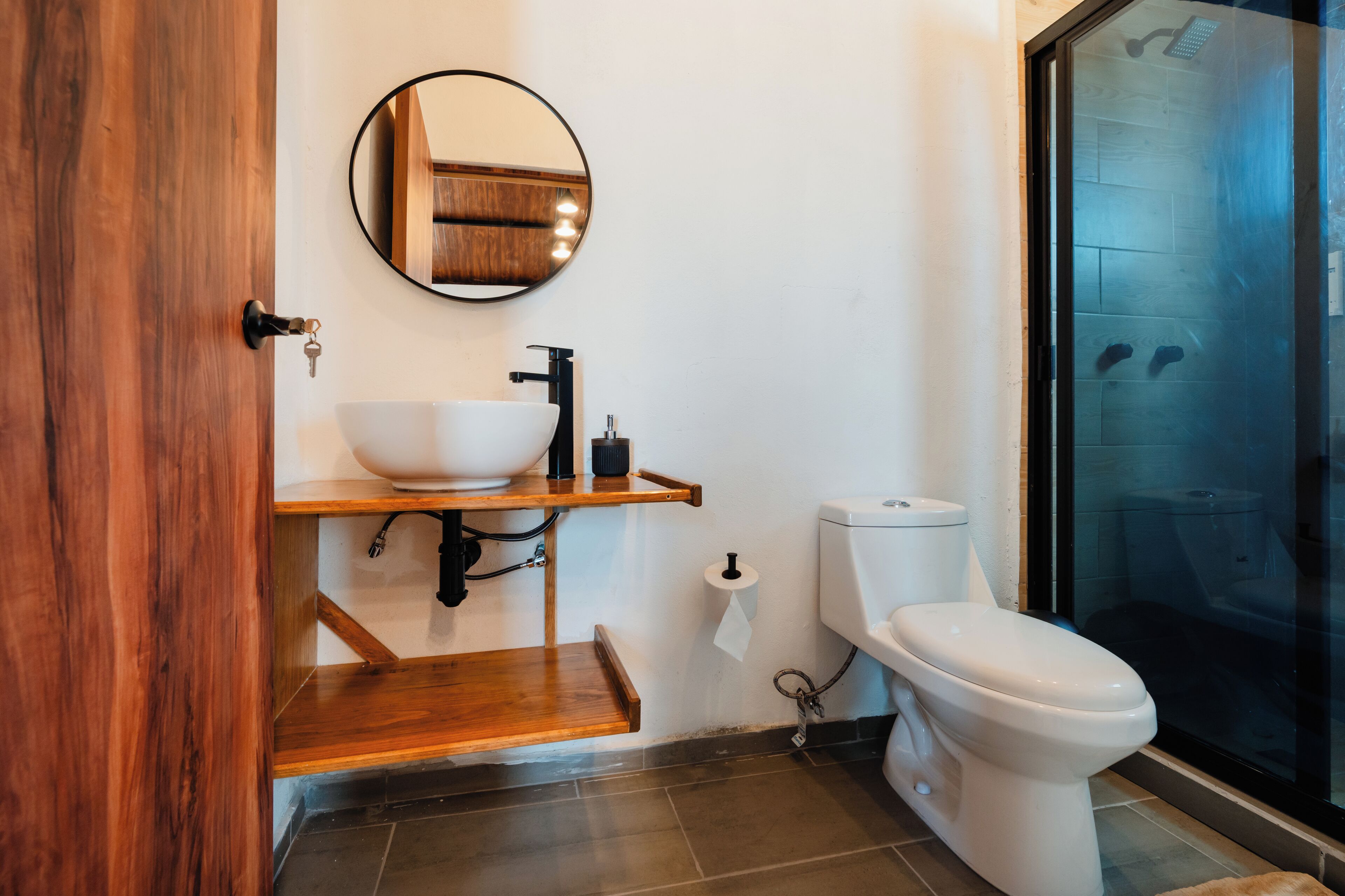 A modern bathroom with a wooden vanity, a round mirror, a white toilet, and a glass shower enclosure.