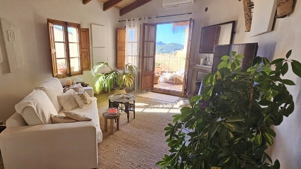 A cozy living room with a sofa, a small table, and a TV. There are plants and a window with curtains. - Single Family Home in Andratx (Andratx)