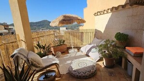 A sunlit patio with a wicker chair, a cushioned bench, a thatched umbrella, and a potted plant. - Single Family Home in Andratx (Andratx)