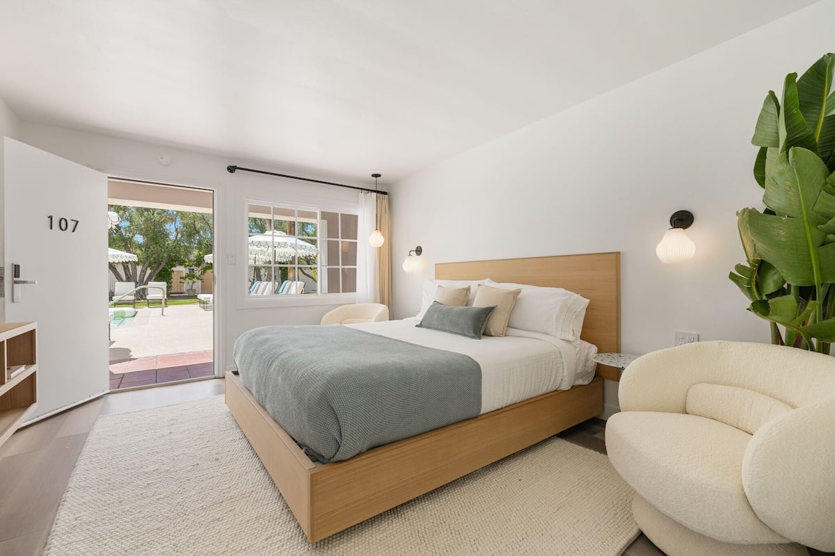 Ein Schlafzimmer mit einem hölzernen Bett, einer weißen Tür mit der Nummer 107, einem weißen Stuhl und einer großen Zimmerpflanze.