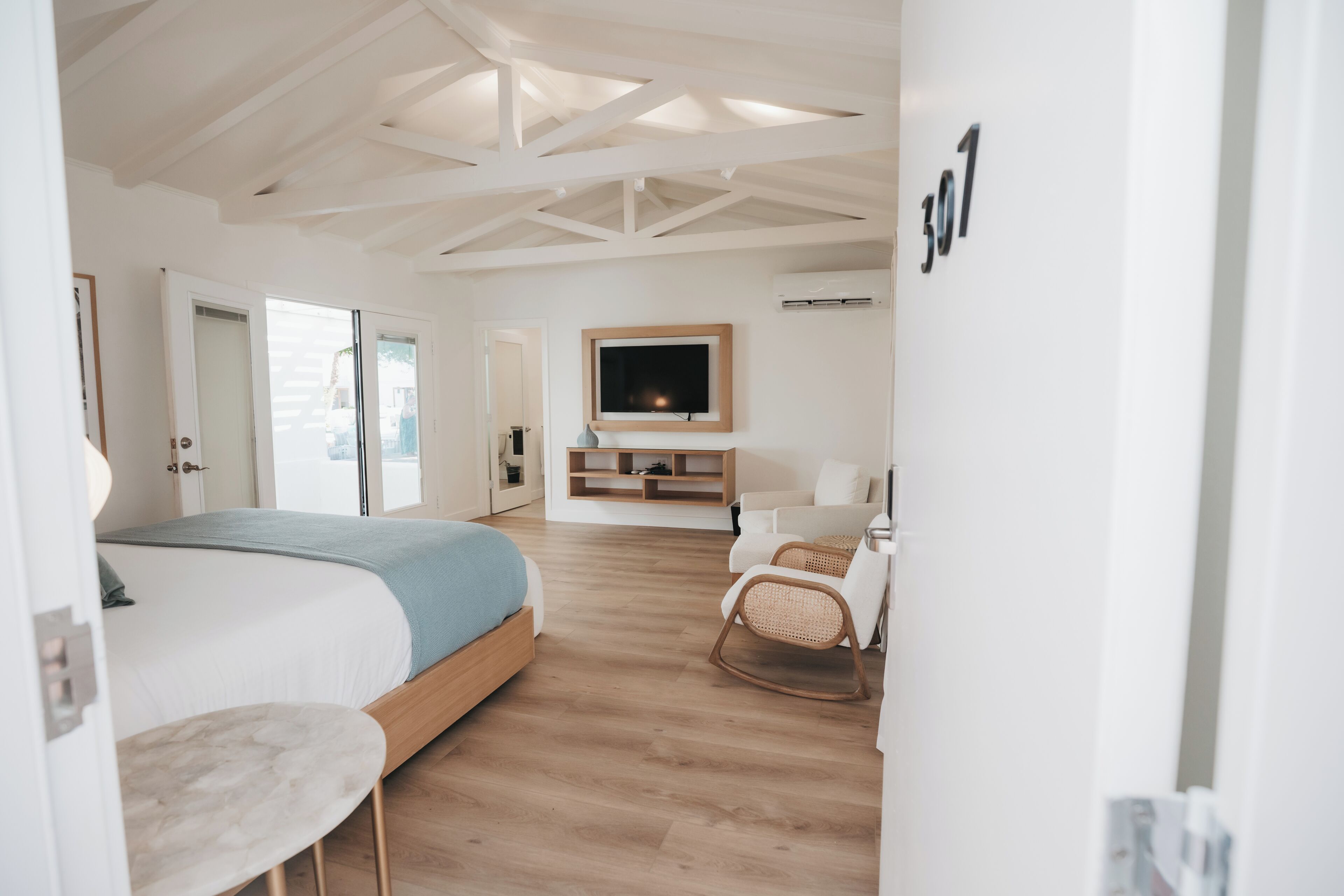 A modern hotel room with a large bed, a TV mounted on the wall, a seating area with a chair, and a wooden floor.
