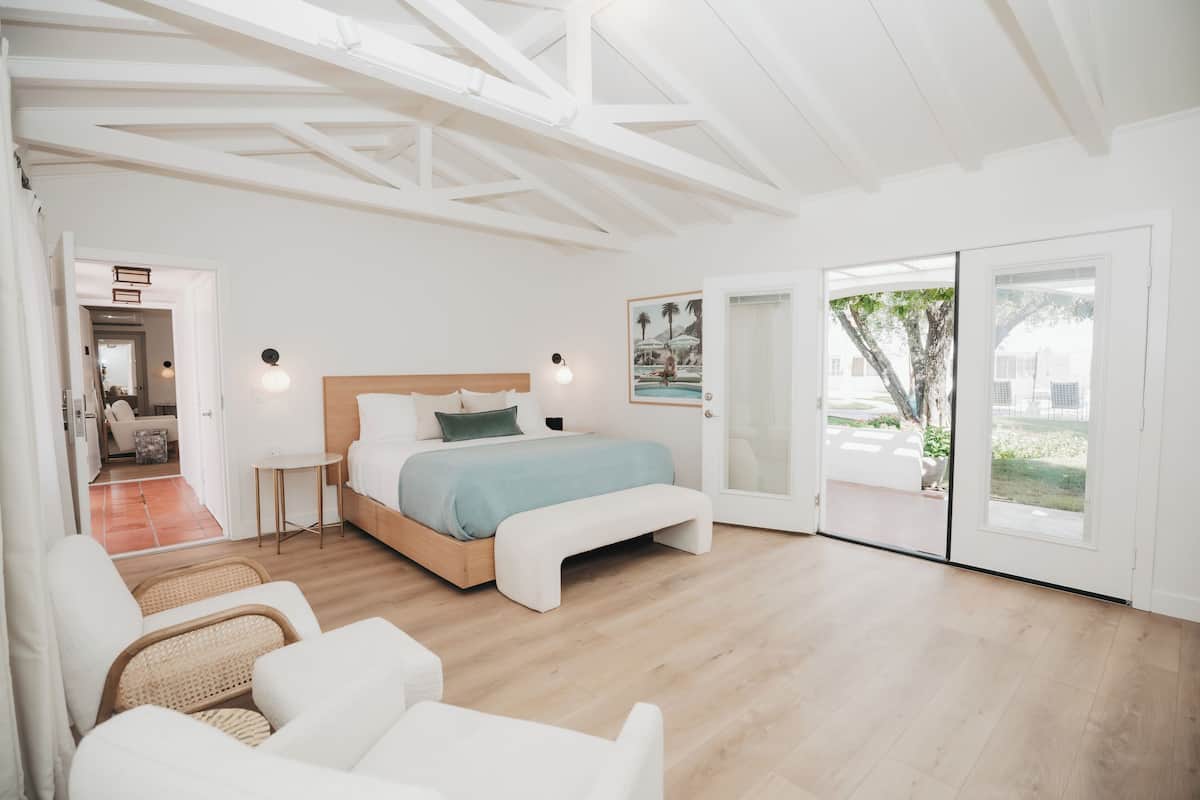 Ein geräumiges Schlafzimmer mit einem großen Bett, einem kleinen Tisch und einer Bank. Eine Tür führt zu einem Balkon mit Blick ins Freie.