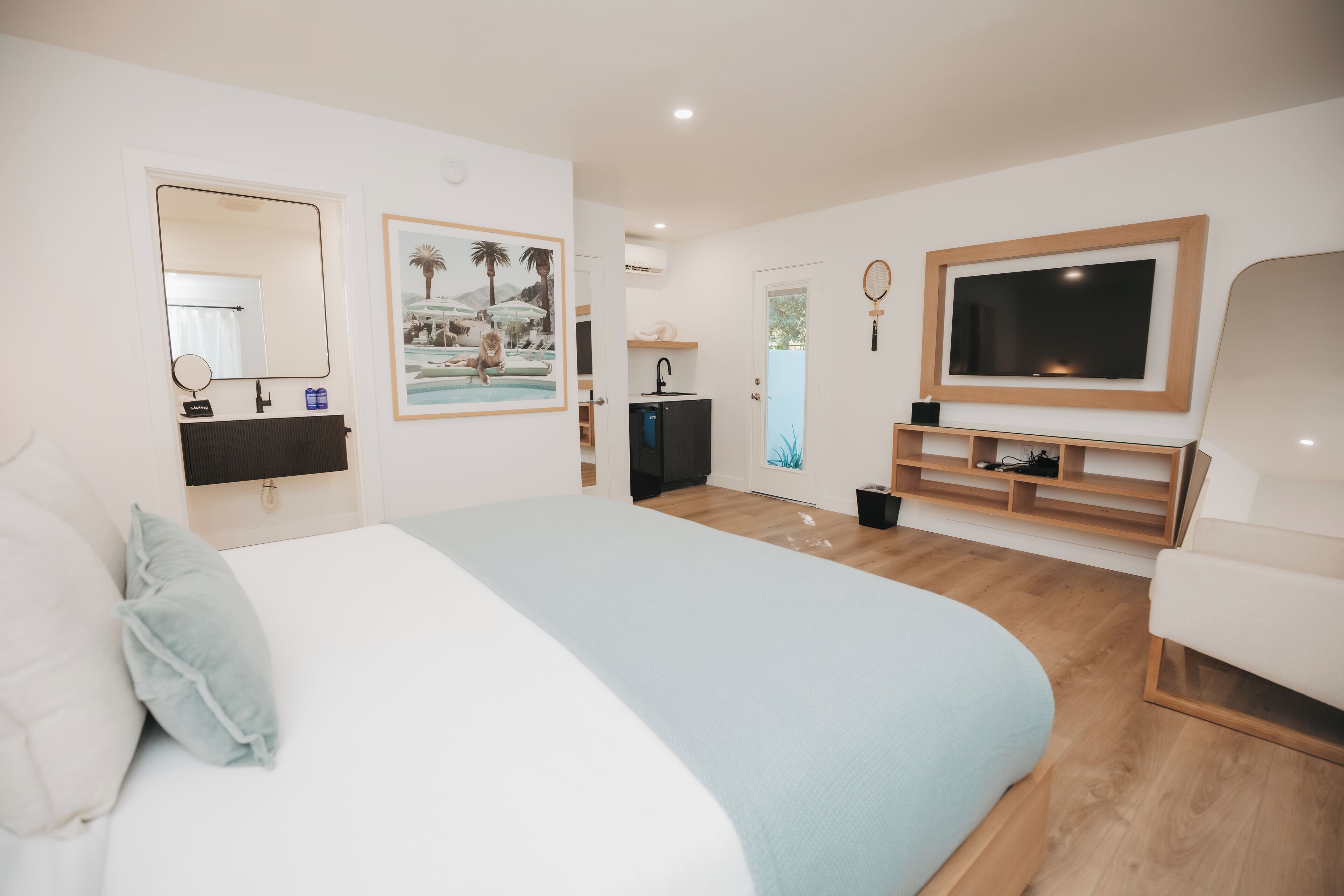 A modern hotel room with a large bed, a TV, a small kitchenette, and a bathroom with a mirror and sink.