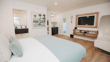 A modern hotel room with a large bed, a TV, a small kitchenette, and a bathroom with a mirror and sink.