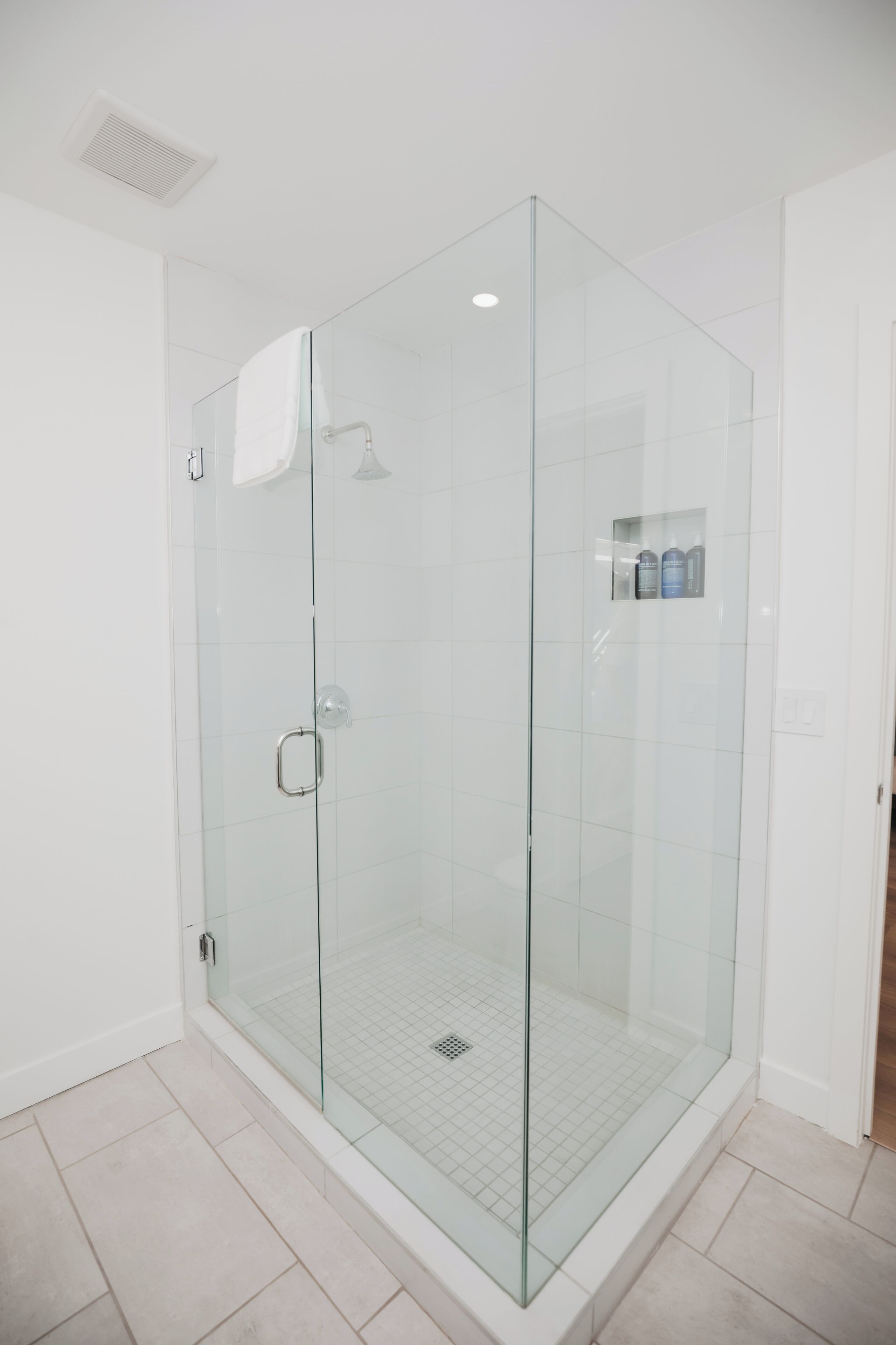 A glass-enclosed shower room with a tiled floor and a small shelf with bottles.