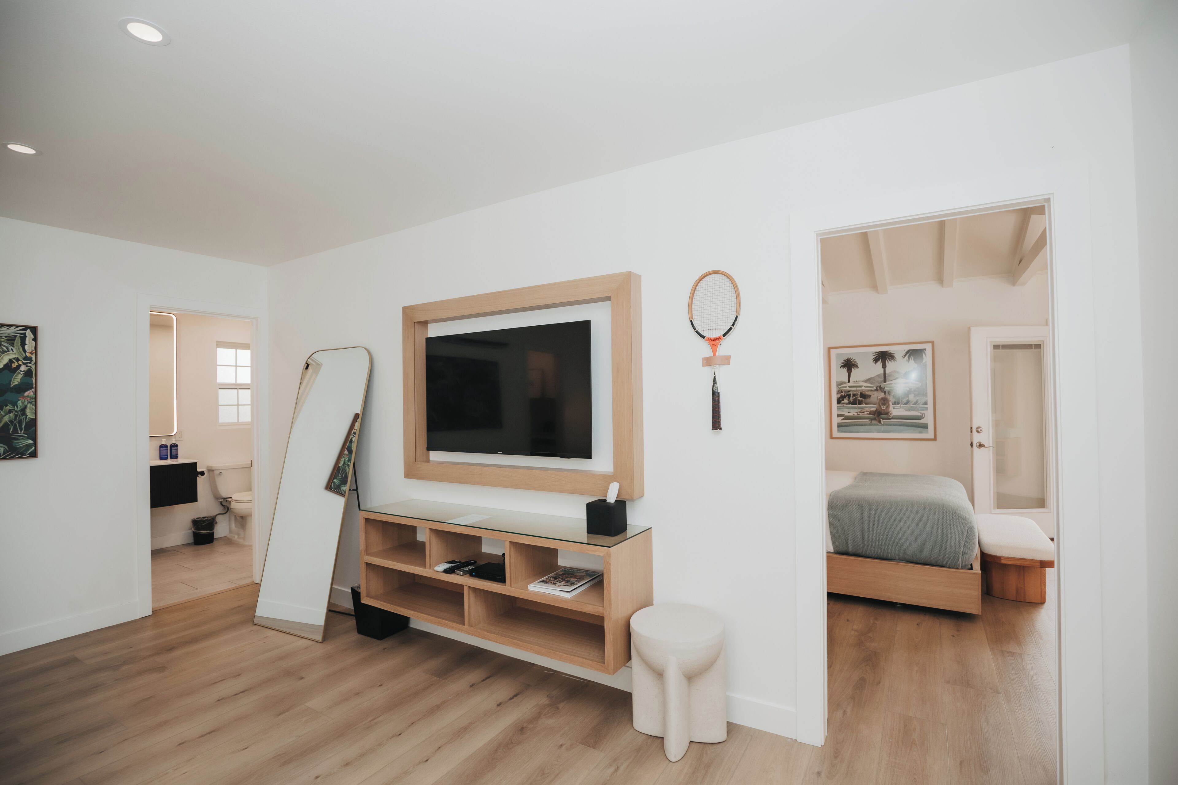 Un dormitorio moderno con televisor, cama, un taburete pequeño y una estantería con libros.