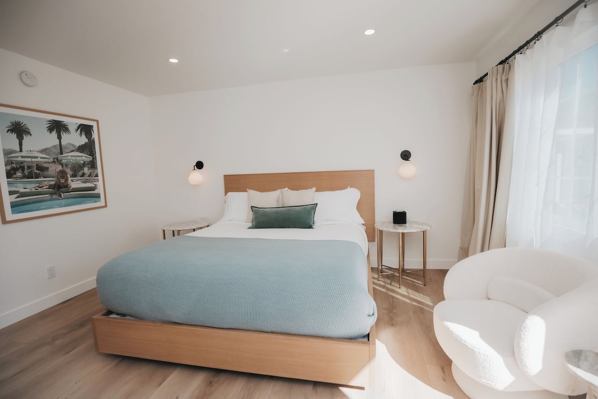 Ein modernes Schlafzimmer mit einem hölzernen Bett, einem kleinen Nachttisch, einem weißen Sessel und einem gerahmten Bild an der Wand.
