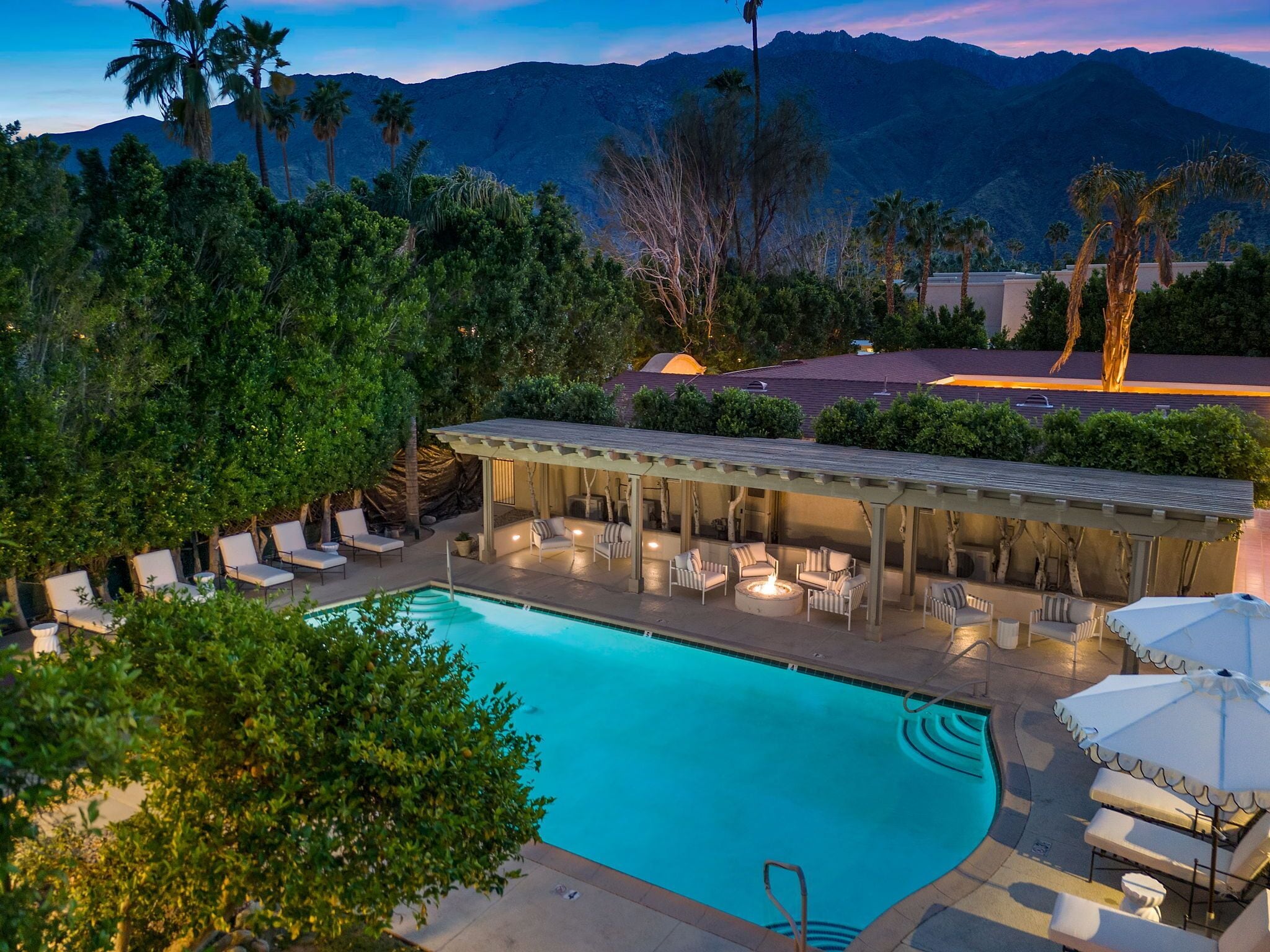A pool area with lounge chairs and a covered seating area, surrounded by trees and mountains in the background.