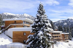 A modern ski resort building with multiple floors, wooden exterior, and a snow-covered mountain backdrop.