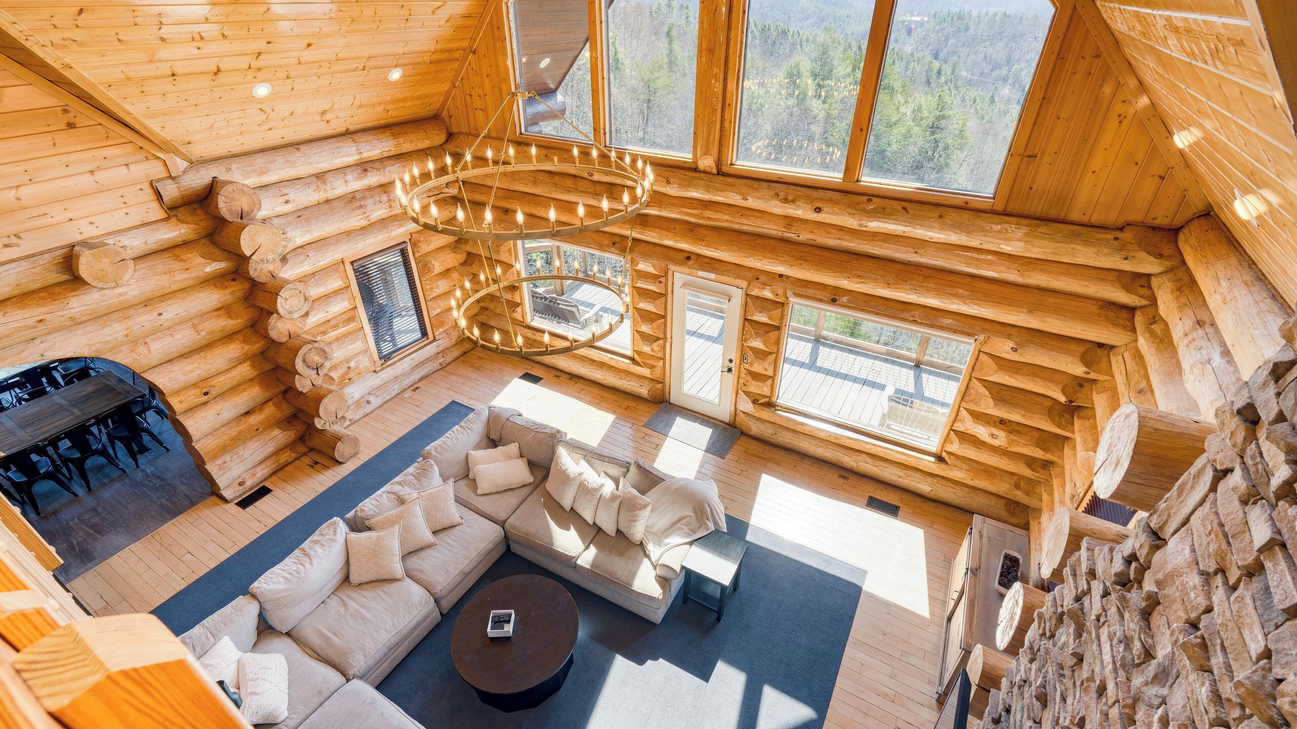 A log cabin interior with a central seating area, a chandelier, and large windows offering a forest view.