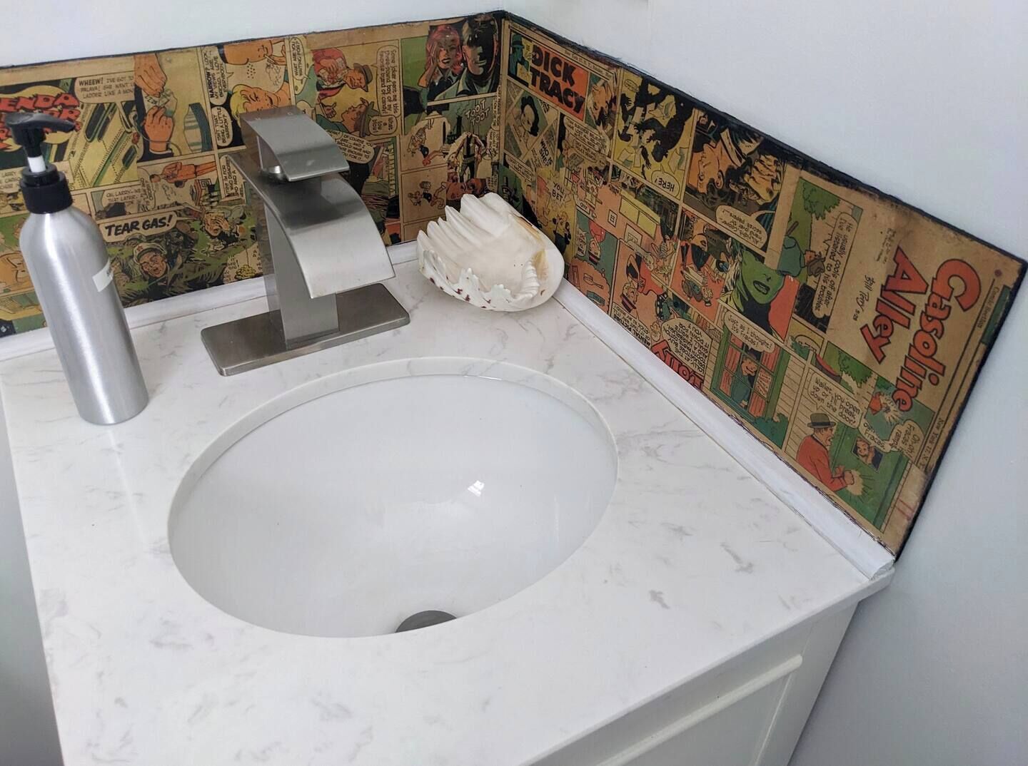 Un baño con lavamanos blanco, grifo plateado y dispensador de jabón. La pared presenta un mural con historietas.