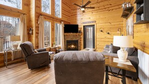 A cozy cabin interior with a fireplace, wooden walls, a ceiling fan, and a television.