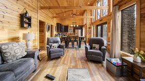A log cabin interior with a wooden ceiling, a dining area, two armchairs, a sofa, and a large window with a stone wall view.