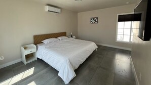 Un dormitorio con cama, mesita de noche, equipo de aire acondicionado, ventana y una obra de arte colgada en la pared.