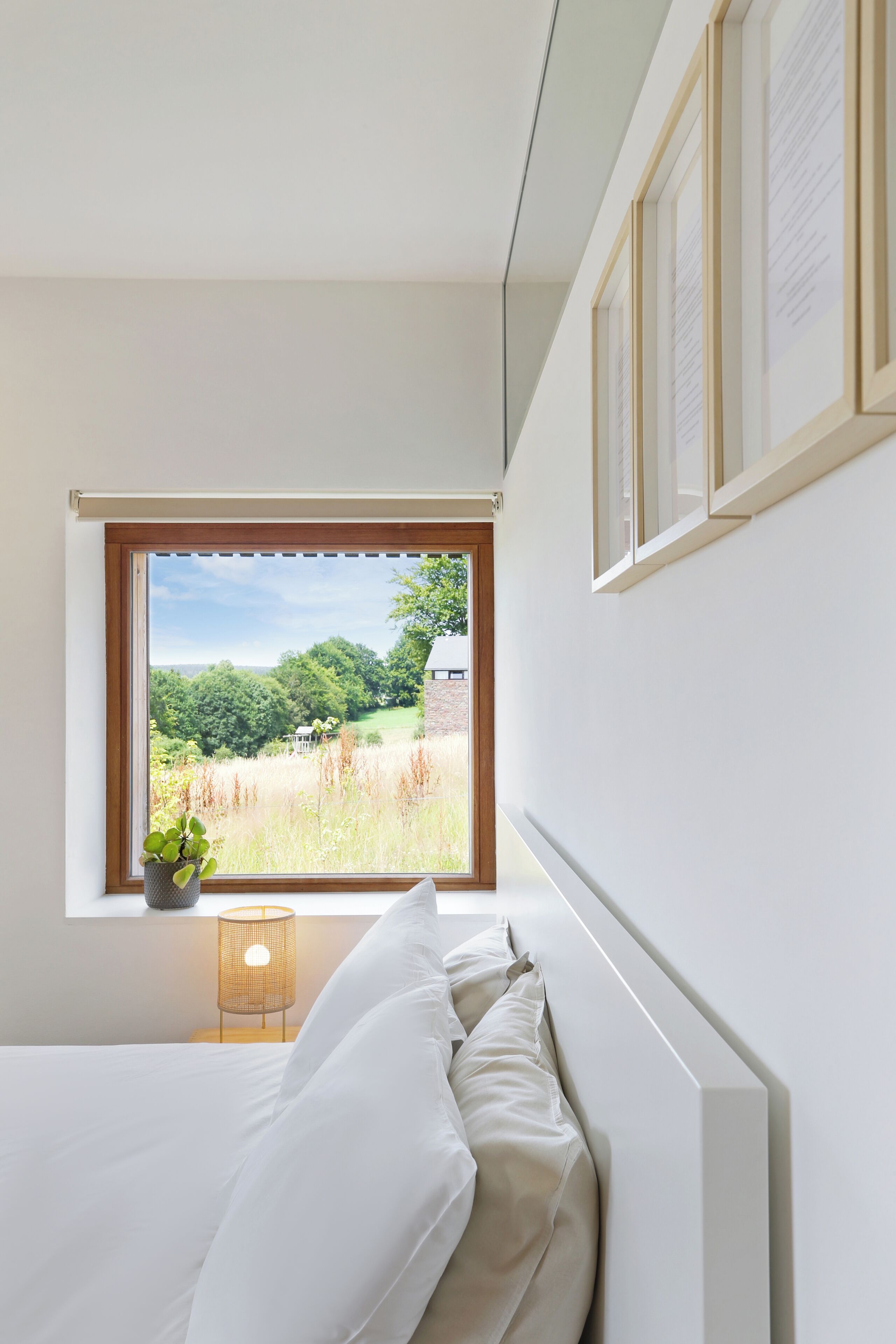 A bedroom with a large window, a bed, a bedside lamp, and a framed notice on the wall.