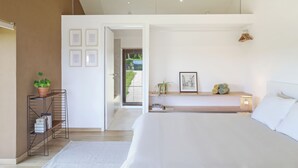 A modern bedroom with a large bed, a bedside table with a lamp, a small shelf with books and a plant, and a view of the outdoors through an open door. - Le pré de Régine et Joseph (Malmedy)