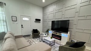 A living room with a beige sofa, a green armchair, a coffee table, and a flat-screen TV mounted on a white wall with geometric paneling.