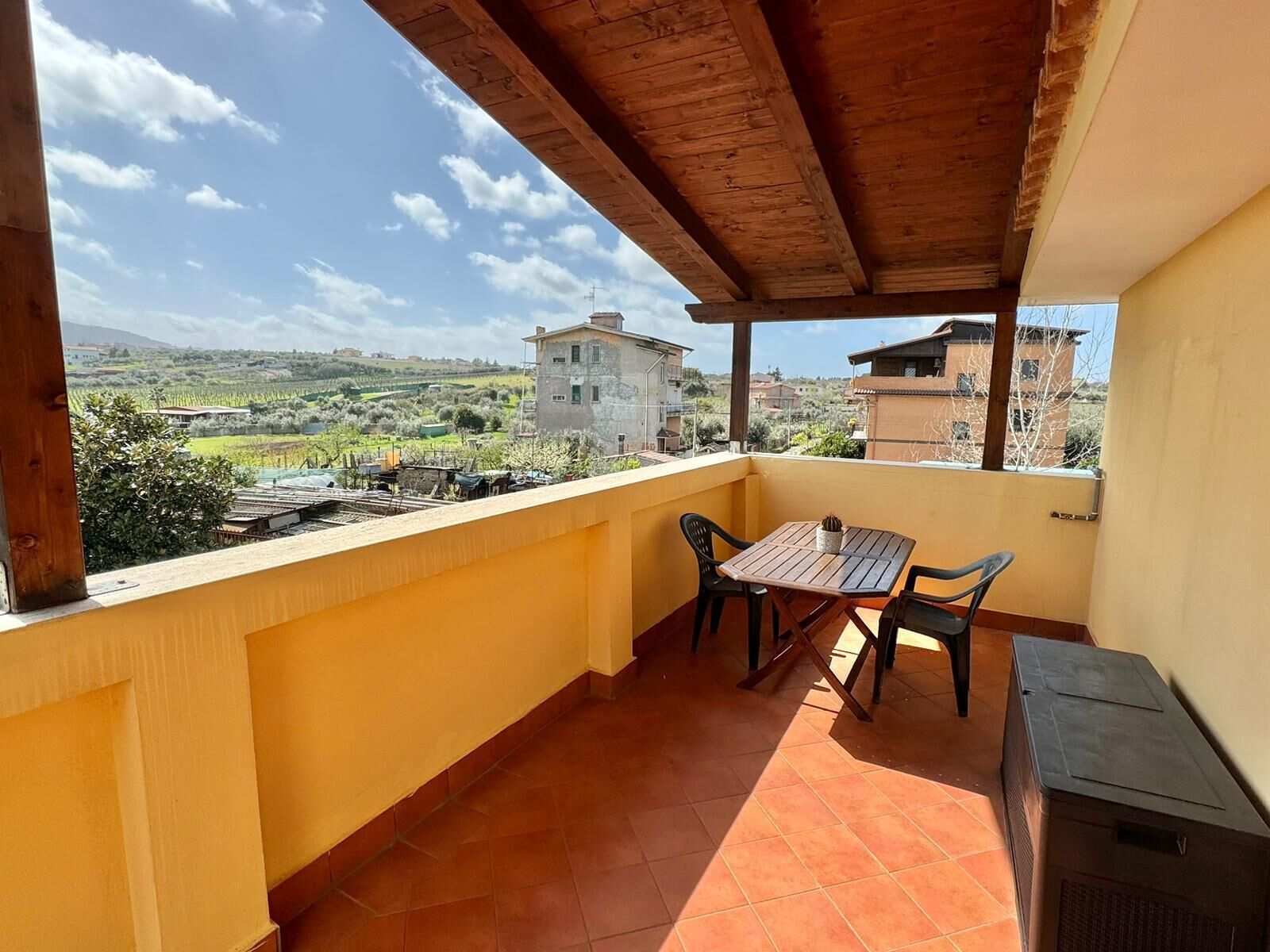 A balcony with a wooden table and chairs, a view of a rural landscape, and a building in the background.