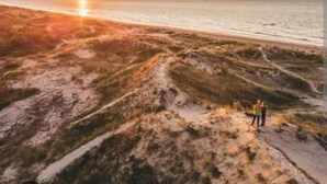 Una playa con un atardecer de fondo y dos personas de pie en la arena.
