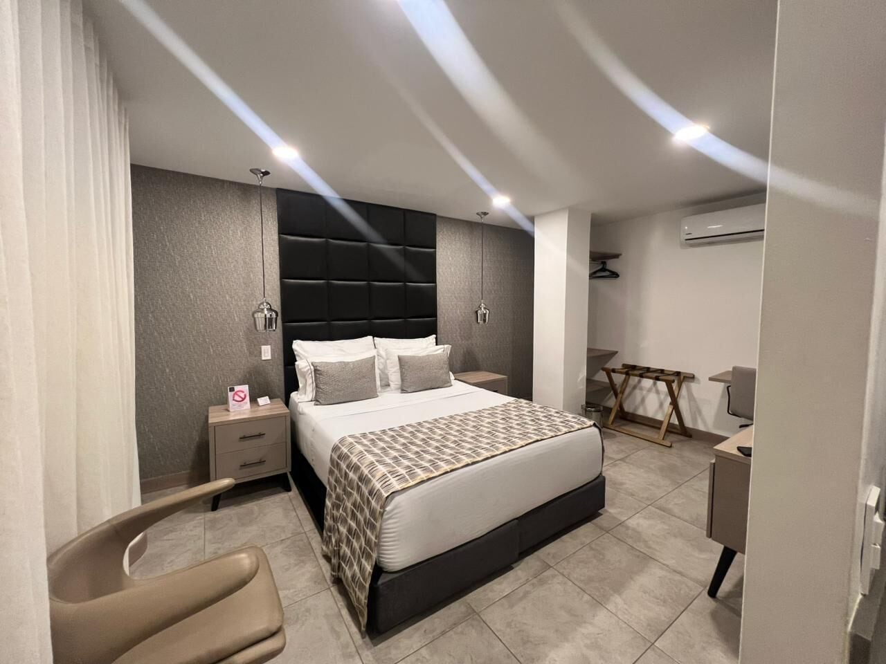 A modern bedroom with a bed, bedside tables, a chair, a desk, and an air conditioning unit.