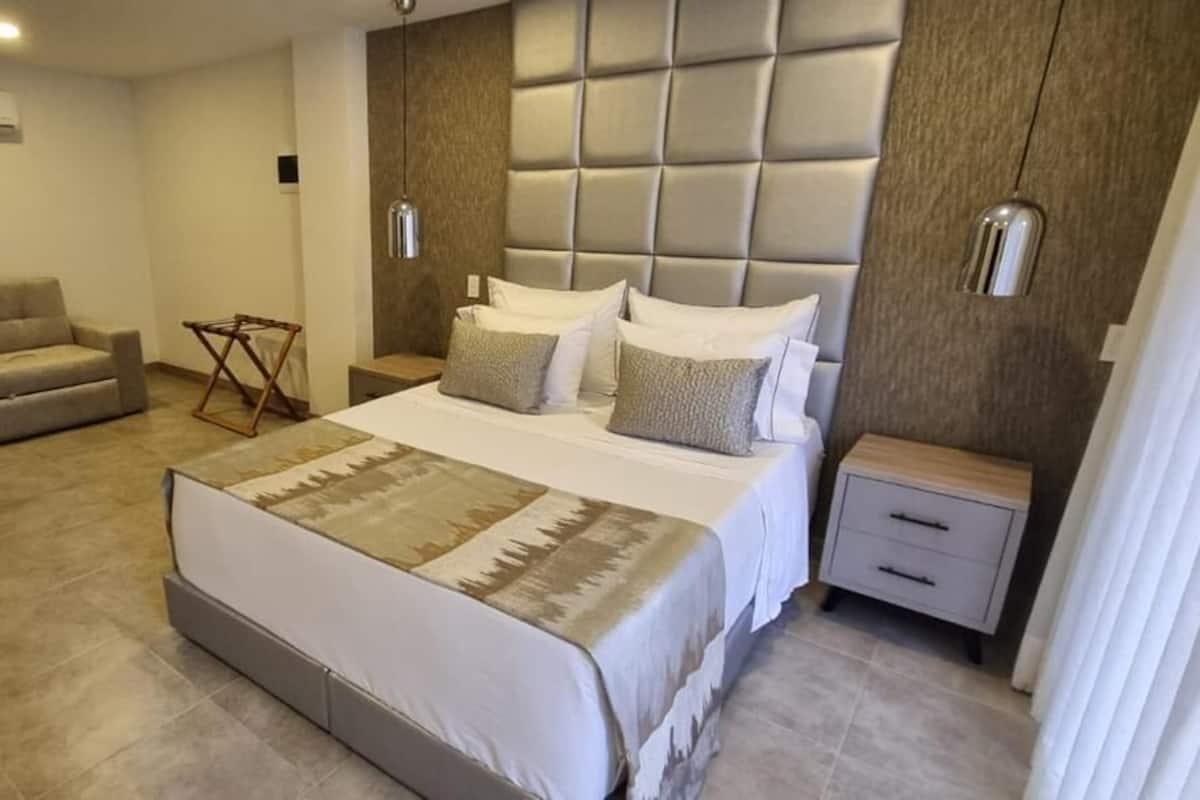 A bedroom with a large bed, a sofa, a nightstand, and a wall-mounted air conditioning unit.