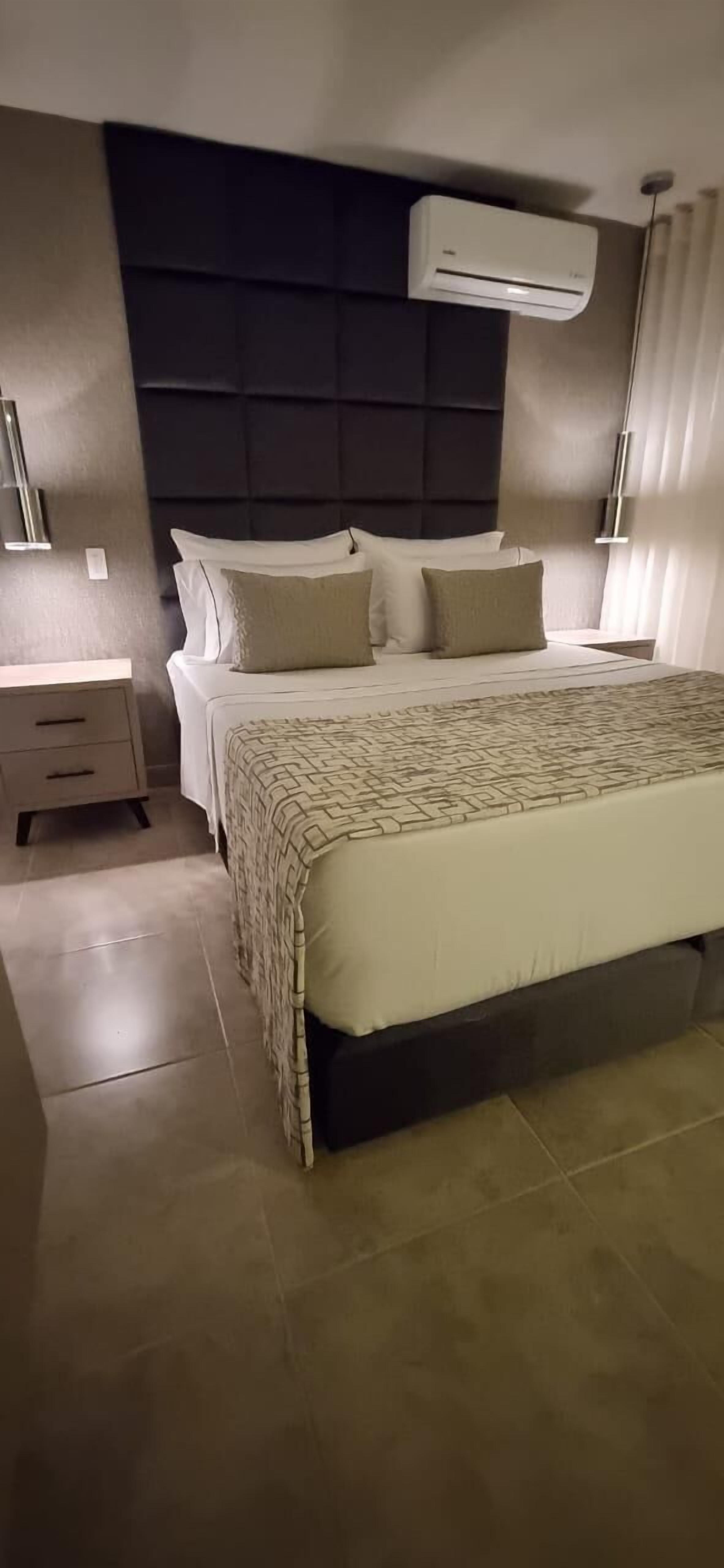 Une chambre d’hôtel moderne avec un grand lit, une tête de lit, un climatiseur, une table de chevet et une lampe.