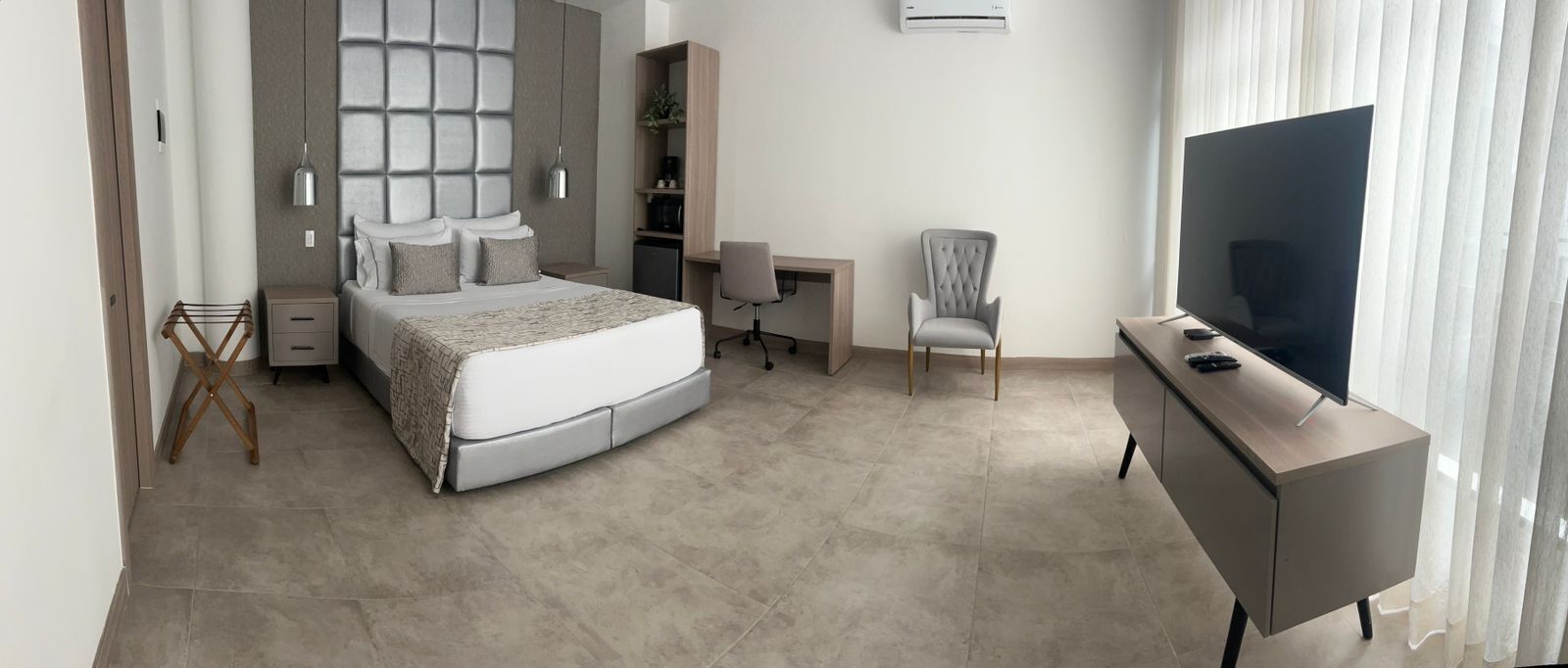 A modern hotel room with a bed, desk, chair, TV, and air conditioning unit.