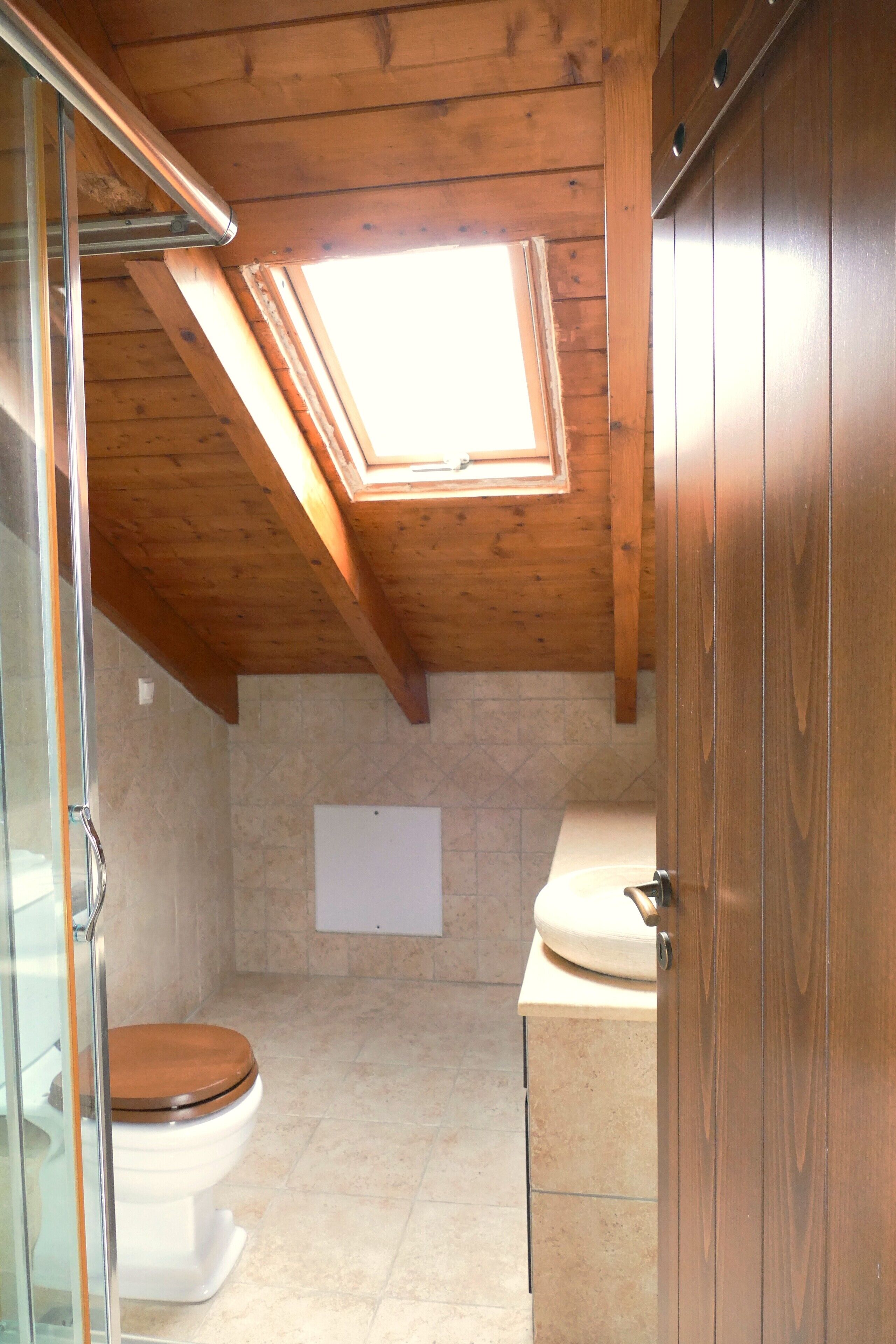 Une petite salle de bain aux dimensions réduites, dotée d’un puits de lumière, de poutres en bois au plafond et de toilettes blanches.