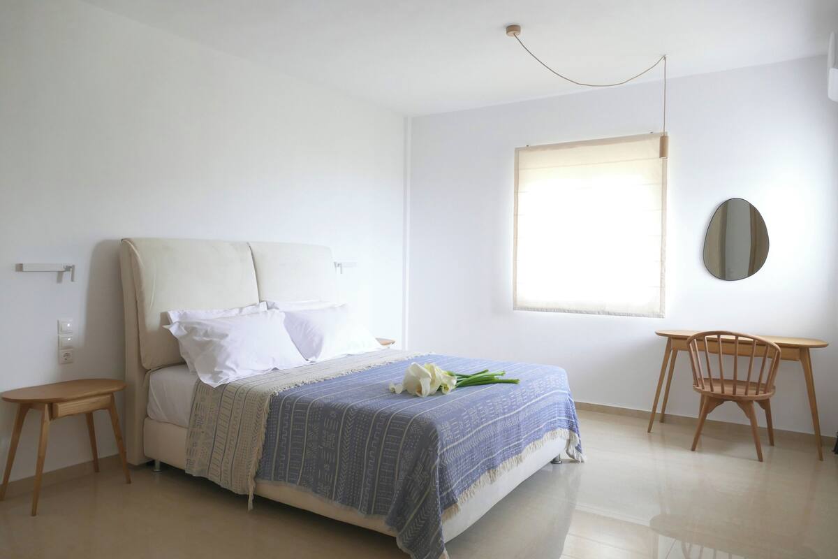 A bedroom with a bed, a wooden chair, a small table, and a mirror on the wall.