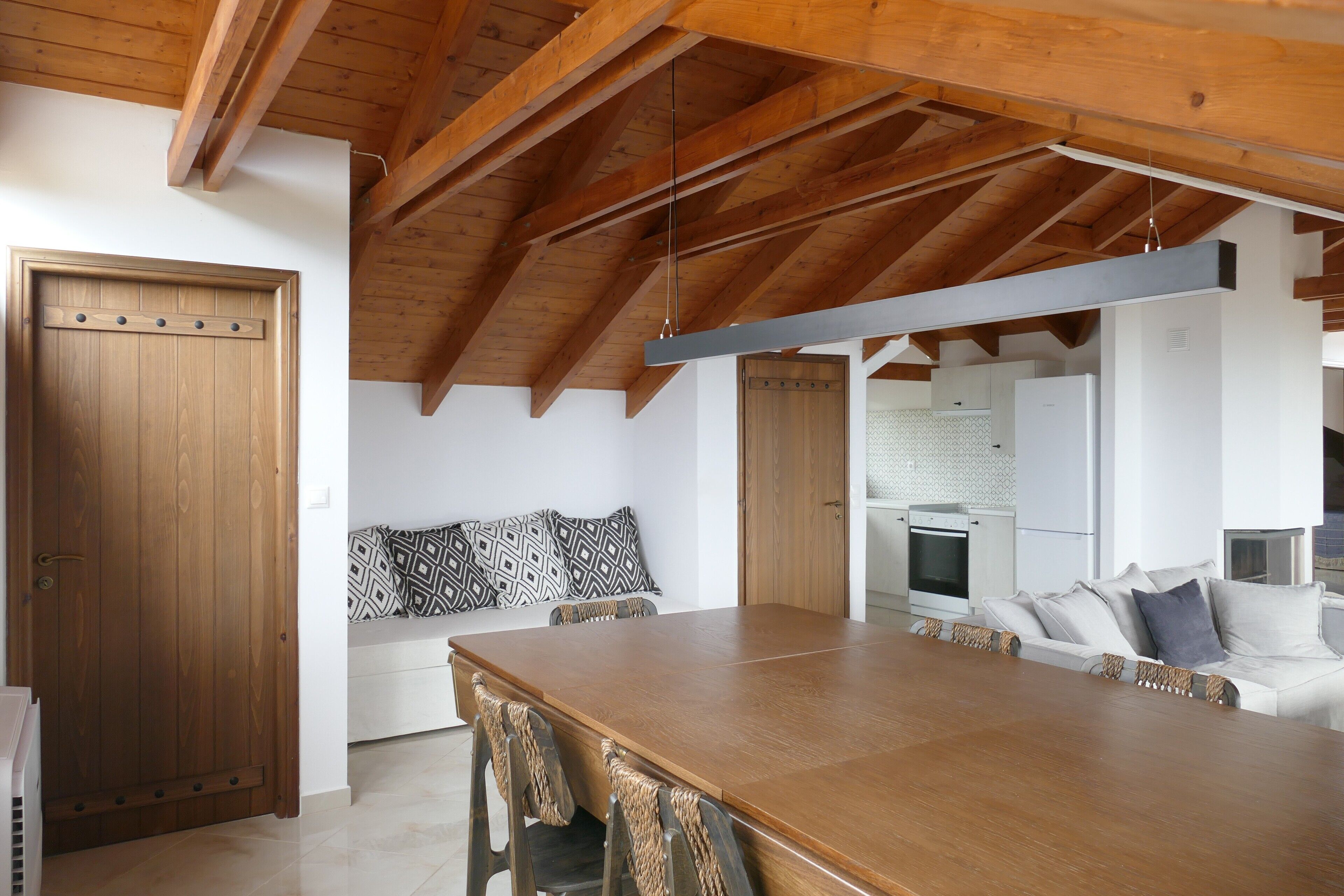 A spacious dining area with a wooden table and chairs, a kitchen in the background, and a high ceiling with exposed beams.