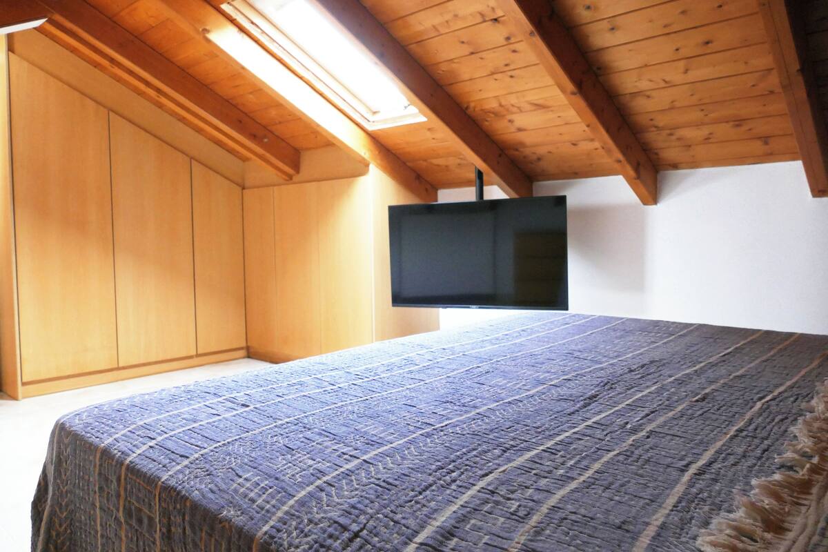 A bedroom with a wooden ceiling, a bed with a patterned bedspread, a wardrobe, and a flat-screen TV mounted on the wall.