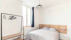 Un dormitorio con cama, cabecera de madera, radiador, ventana con cortinas, lámpara colgante y estante de madera.