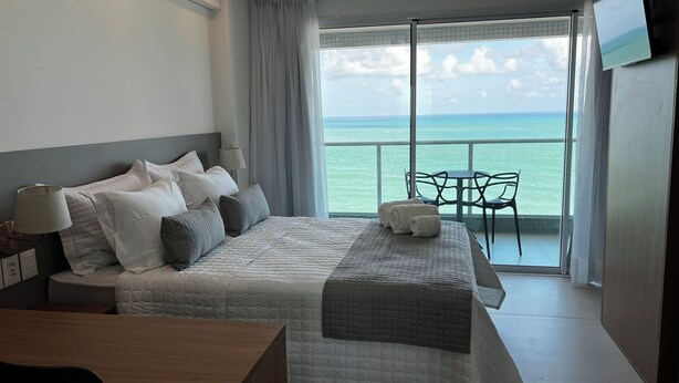 Ein Hotelzimmer mit einem großen Bett, einem Schreibtisch, einem Sessel, einem Fernseher und einem Balkon mit Meerblick.