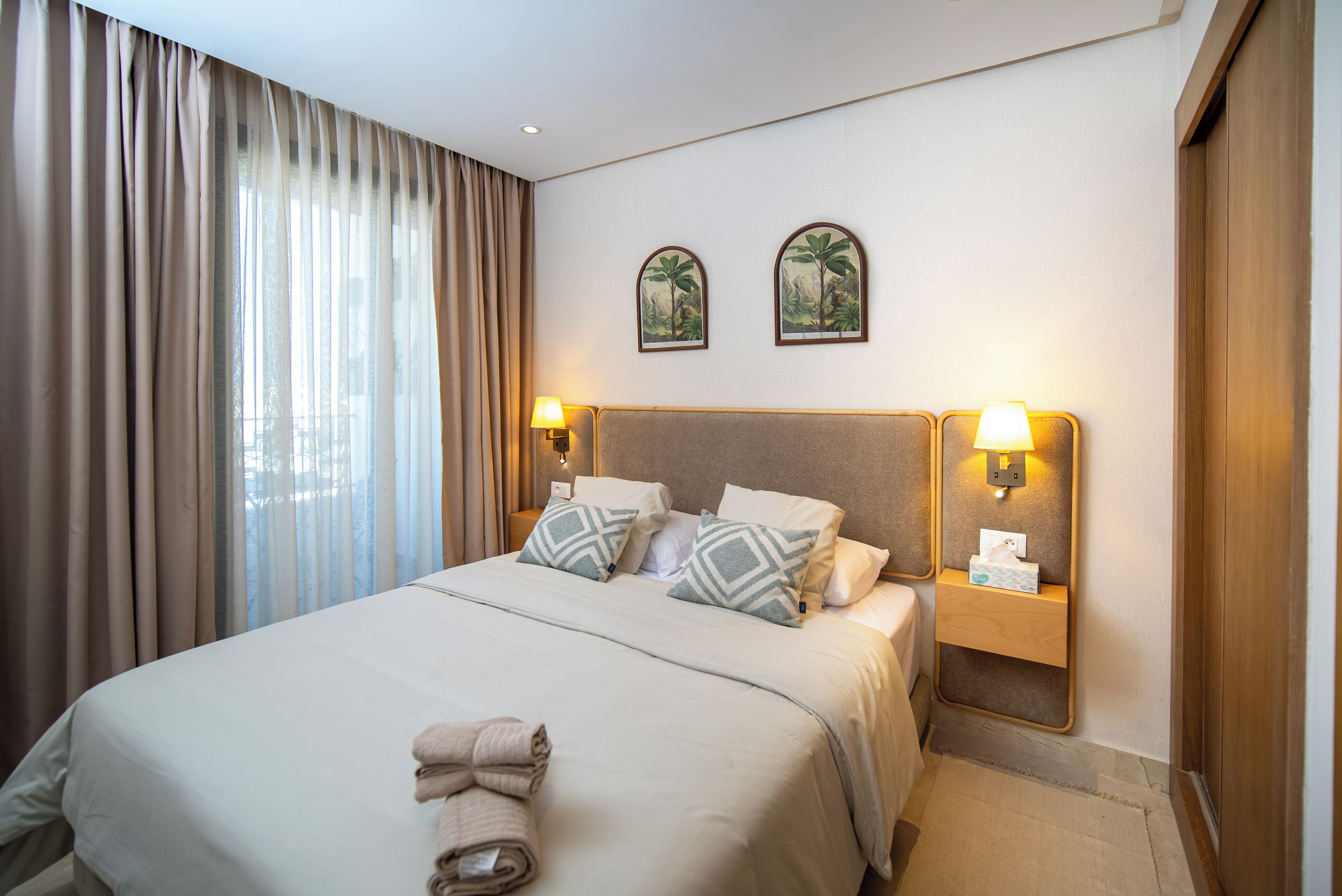 Una habitación de hotel con una cama grande, dos lámparas fijadas a la pared y un ventanal con cortinas.