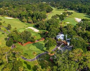 Vue aérienne d’un terrain de golf comportant plusieurs trous, entouré d’une forêt dense et d’un espace de stationnement.