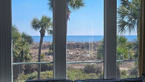 Blick aus einem Fenster mit einer Couch im Vordergrund auf einen Strand mit Palmen und einem klaren Himmel.