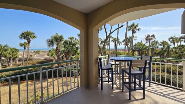 Ein Balkon mit Tisch und Stühlen, der auf einen Strand mit Palmen und den Ozean blickt.