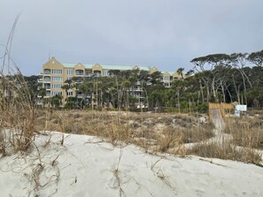 A beachfront hotel with multiple balconies, surrounded by sand dunes and sparse vegetation.
