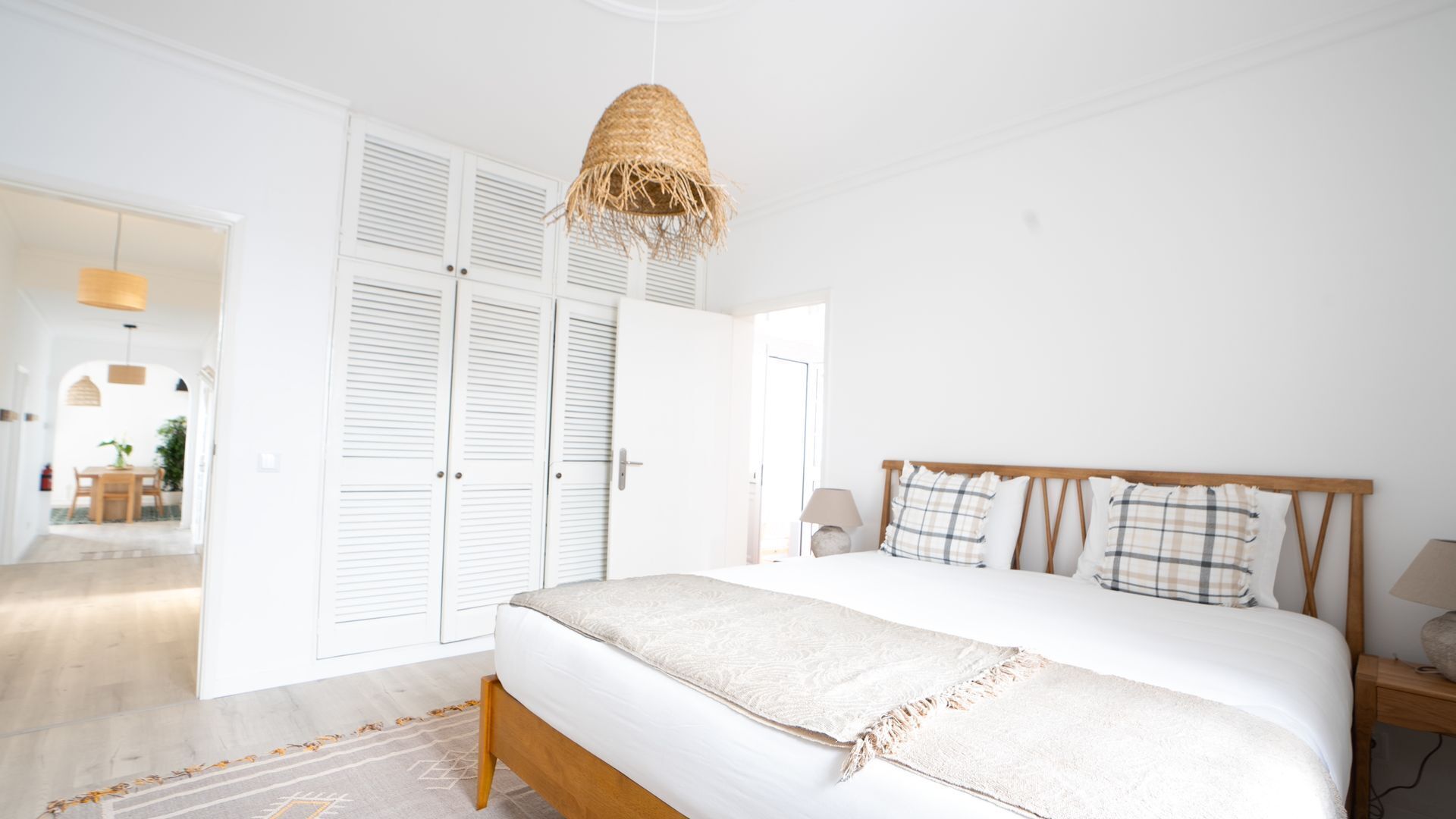 Une chambre à coucher avec un grand lit, une tête de lit en bois, un luminaire suspendu et une porte donnant sur une autre pièce.