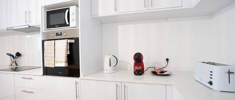 Una cocina moderna con armarios blancos, microondas integrado, hervidor, cafetera y tostadora.