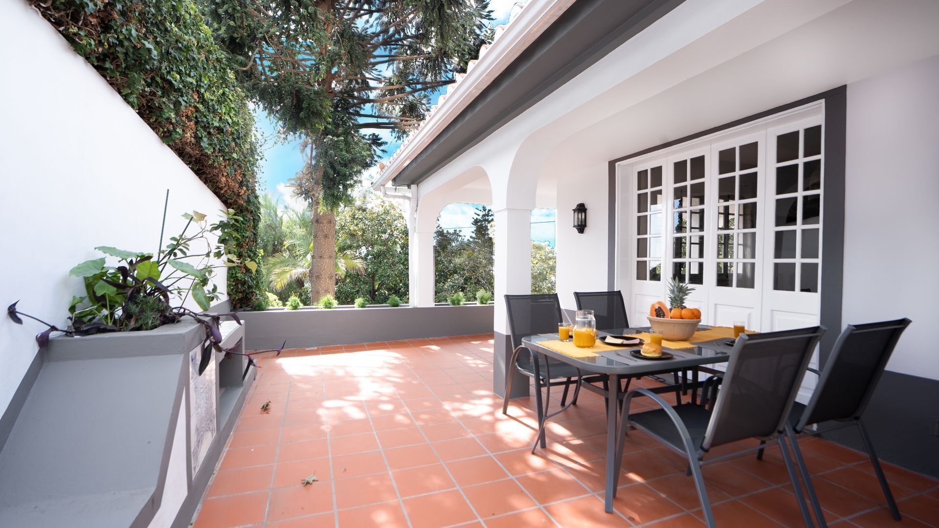 A patio with a table set for breakfast, surrounded by chairs, with a view of greenery and a white door.