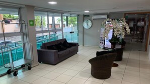A reception area with a sofa, wicker chair, and a cart near large windows. - Melhores Flats - Sossego frente ao mar (João Pessoa)