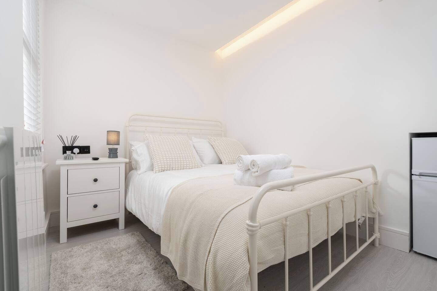 A bedroom with a white bed, a nightstand, a lamp, and a refrigerator.