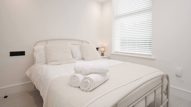 A neatly made bed with white bedding and pillows, a bedside table with a lamp, and a window with blinds. - Near Anfield | SofaBed & Mini Fridge | Parking (Liverpool)
