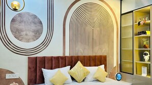 A modern bedroom with a large bed, a ceiling fan, a wall-mounted shelf with decorative items, and a circular light fixture. - Cozy studio in marvelous Pune with AC, WiFi, Yashvantgad. (Pune)