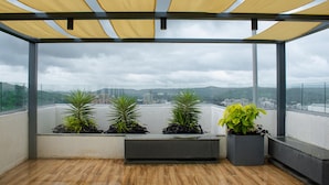 A balcony with a wooden floor, potted plants, and a view of a cityscape. - Cozy studio in marvelous Pune with AC, WiFi, Yashvantgad. (Pune)