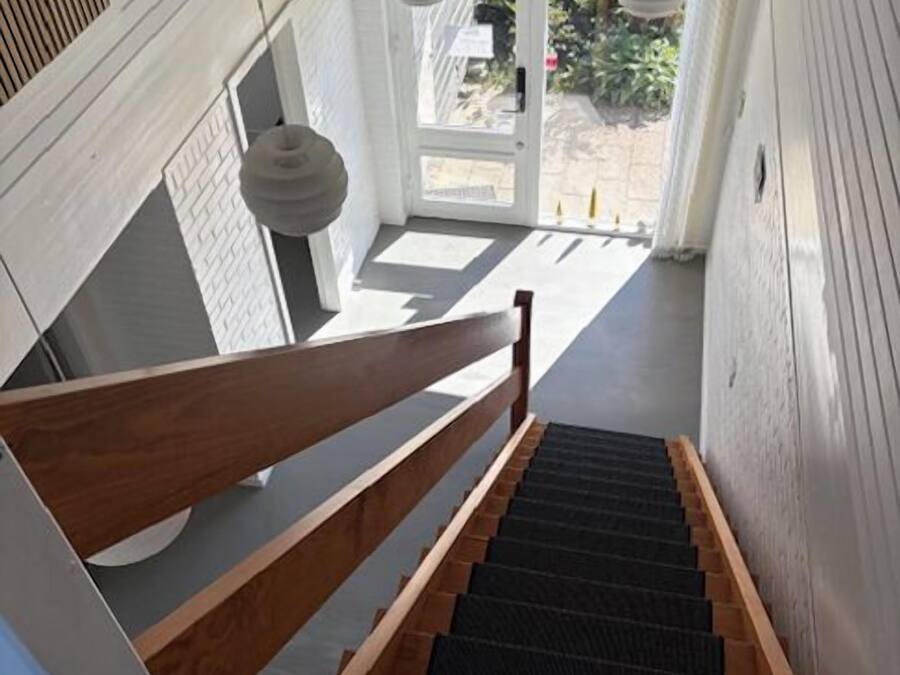 Una escalera de madera con barandilla que lleva a una puerta con vistas al exterior, donde se aprecia vegetación.