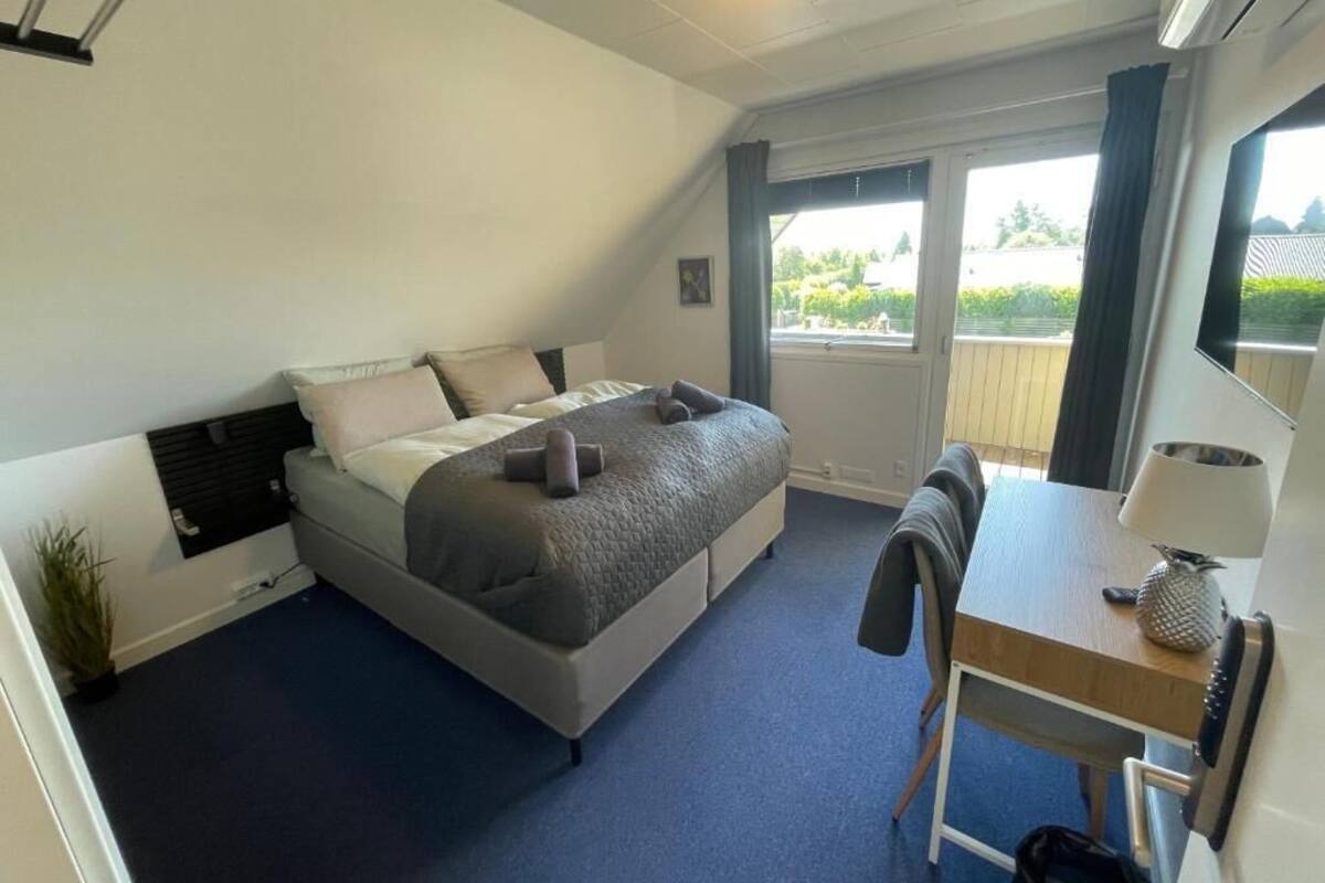 A bedroom with a bed, a desk, a chair, a lamp, and a window with a view of greenery.